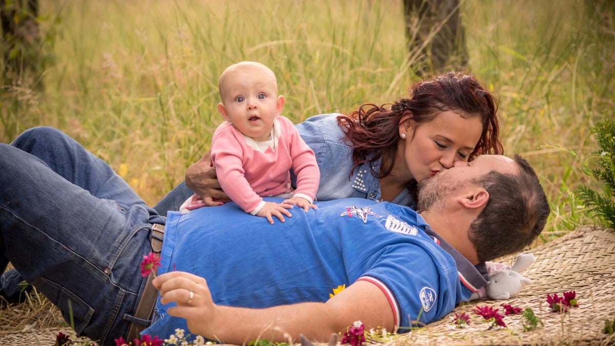 A tender family scene outdoors with parents kissing while their baby lies on the father.